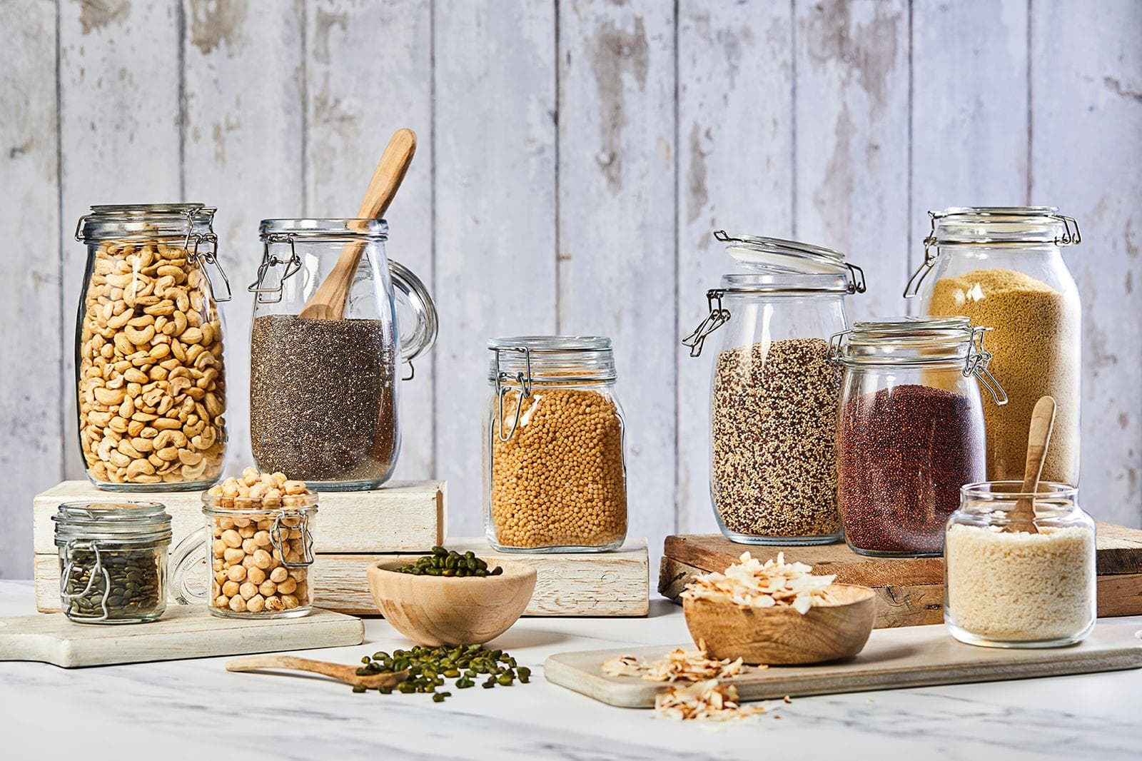 A collection of glass storage jars filled with various dried ingredients including cashews, chia seeds, lentils, quinoa, rice, and pulses, arranged on wooden blocks against a white wooden background.
