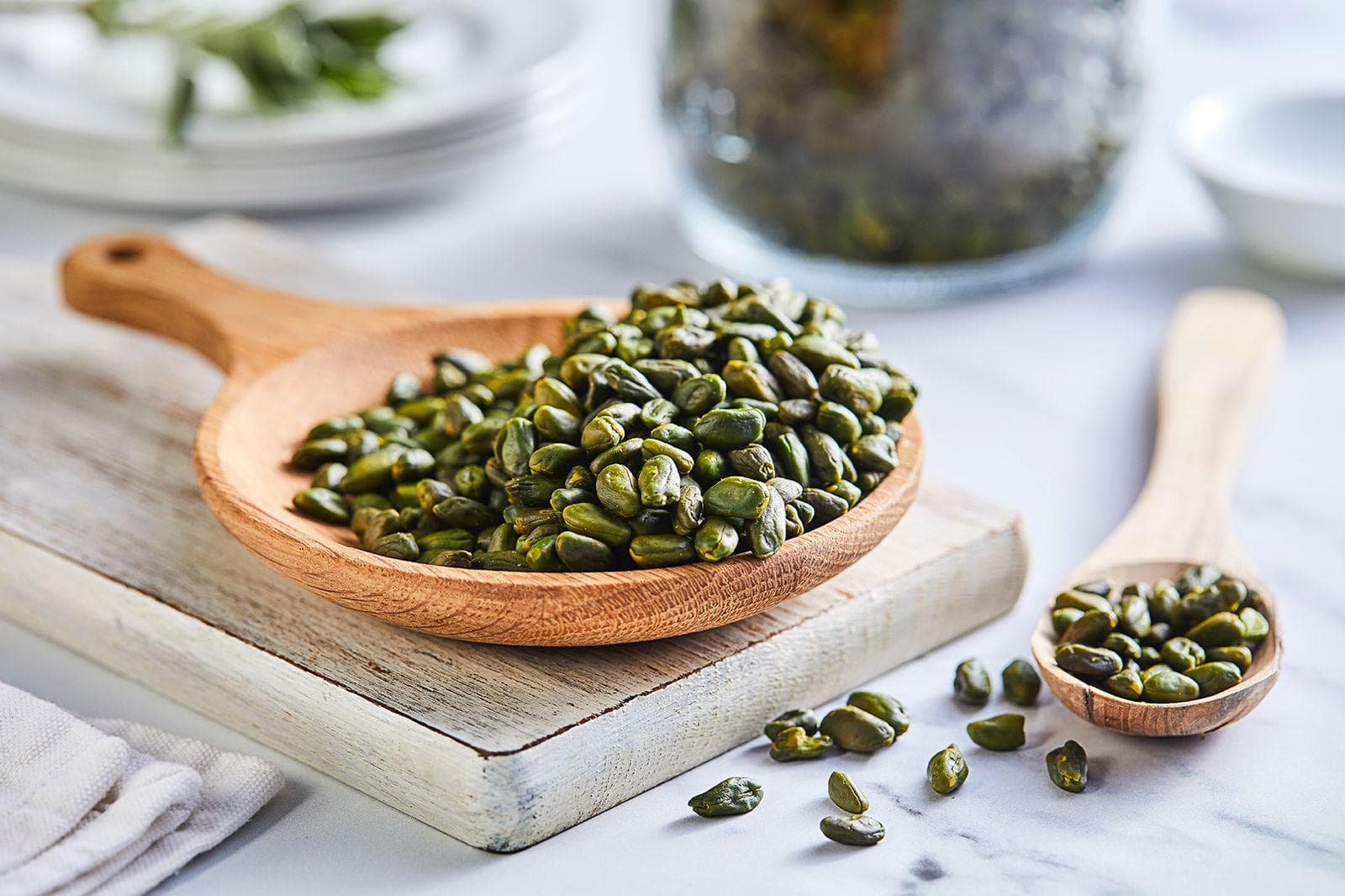 Green pumpkin seeds (pepitas) displayed in wooden spoons on a white marble surface, styled with plates and kitchen linens.
