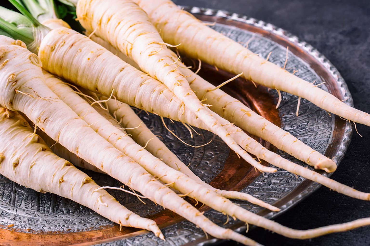 Several fresh white parsnips with roots and soil still attached, arranged on a dark decorative plate with a dark background.