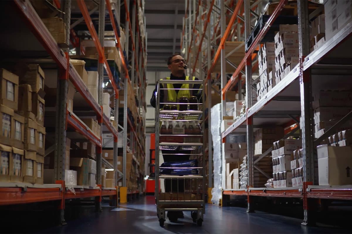 A worker in a high-visibility vest operating a motorised order picker lift truck between tall red and white industrial shelving units in a large warehouse, filled with cardboard boxes and packaged goods.