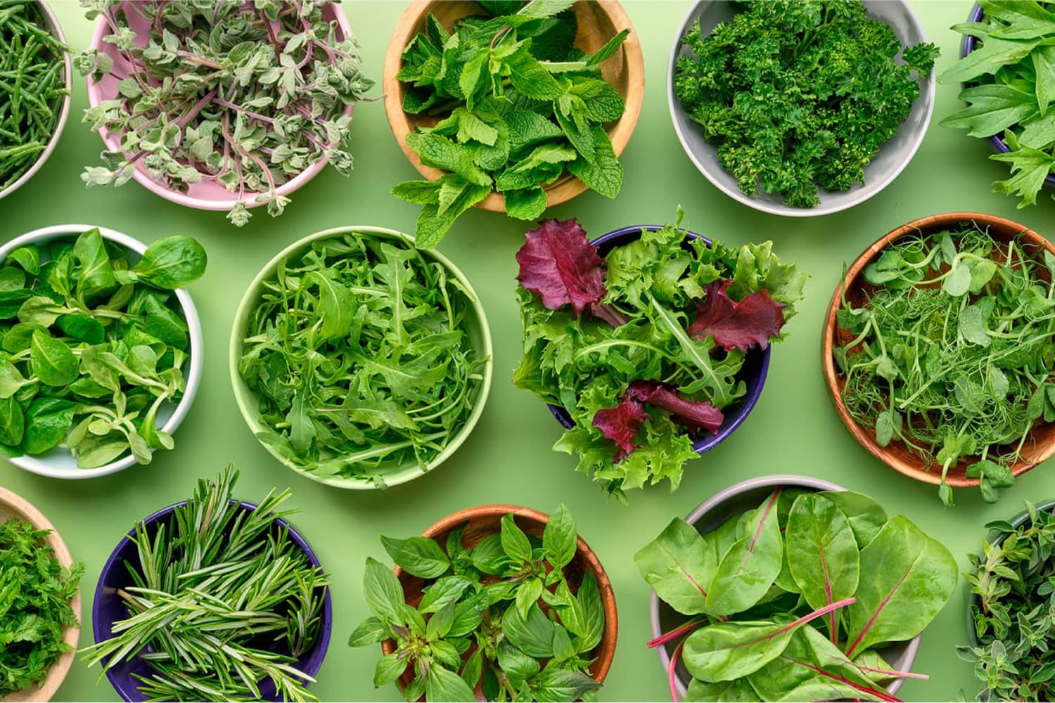 Overhead view of various fresh herbs and salad leaves arranged in colourful bowls on a green background, including mint, parsley, rocket, spinach, and mixed lettuce varieties.