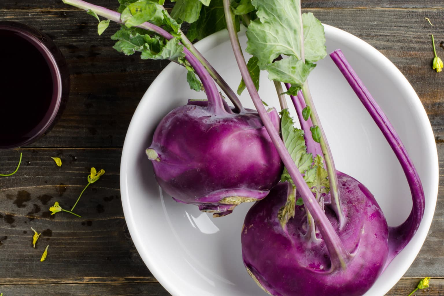 Two fresh purple kohlrabi bulbs with vibrant green leaves and purple stems arranged on a white plate, displayed on a dark wooden surface with small yellow flowers scattered nearby.