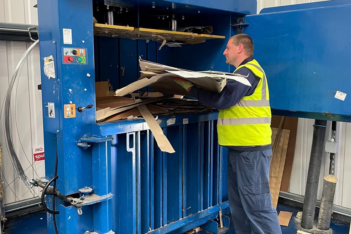 A worker in a high-visibility vest operating a large blue industrial waste compactor machine, loading cardboard and wooden materials into the press chamber.