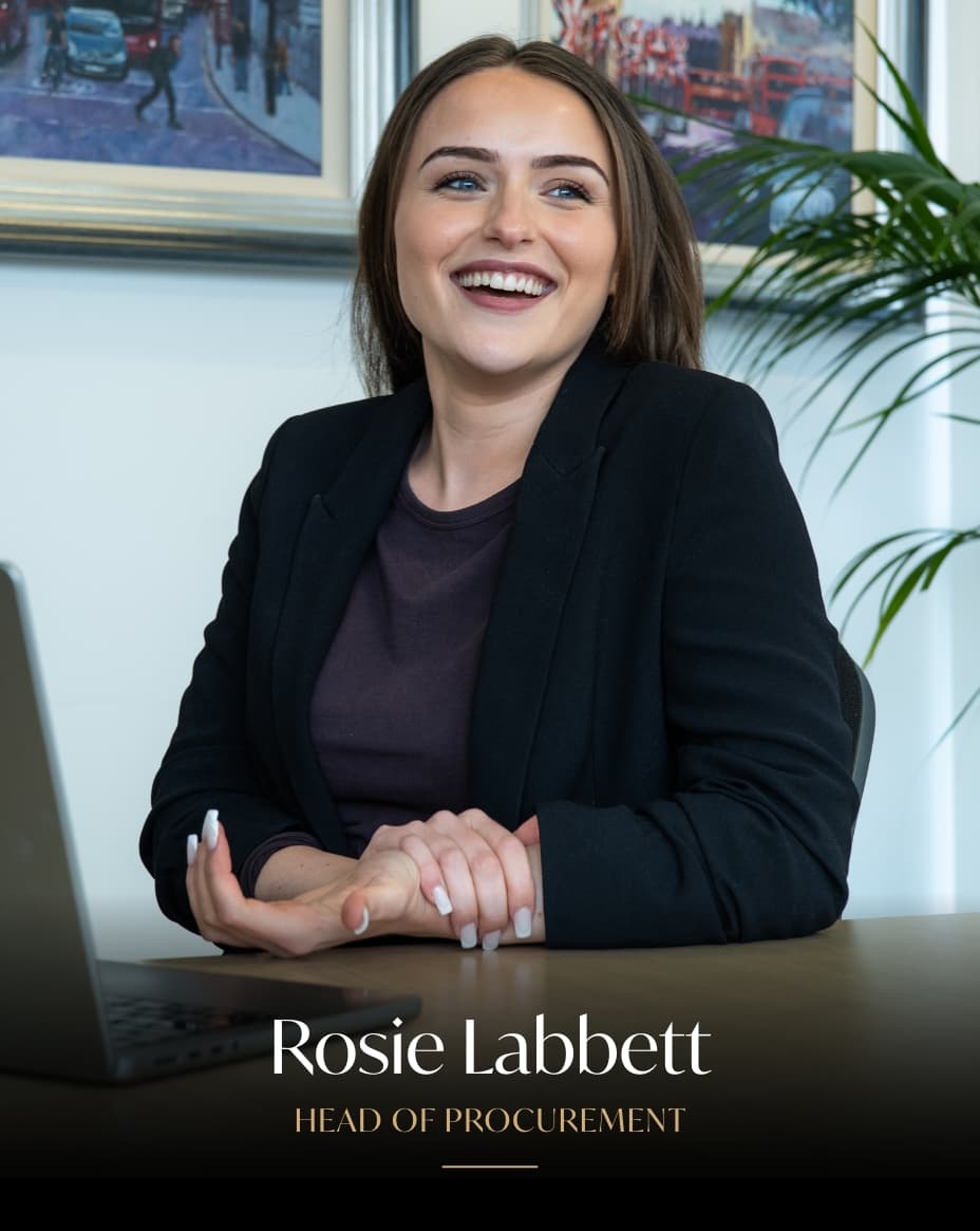 Professional headshot of Rosie Labbett, Head of Procurement, smiling at desk in office setting with plants and framed artwork in background.