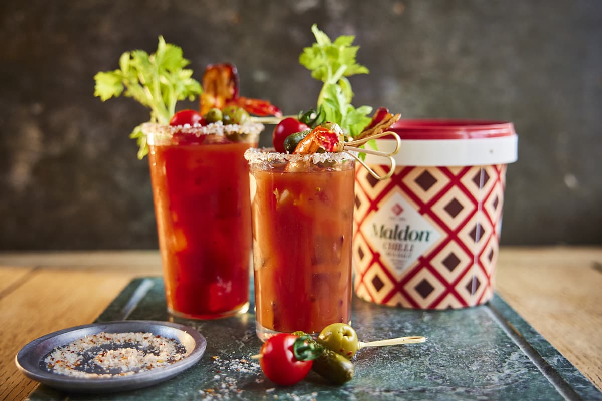 Two red cocktails with celery garnish and seasoned rims alongside a container of Maldon salt, decorated with cocktail ingredients including olives and peppers on a dark slate surface.