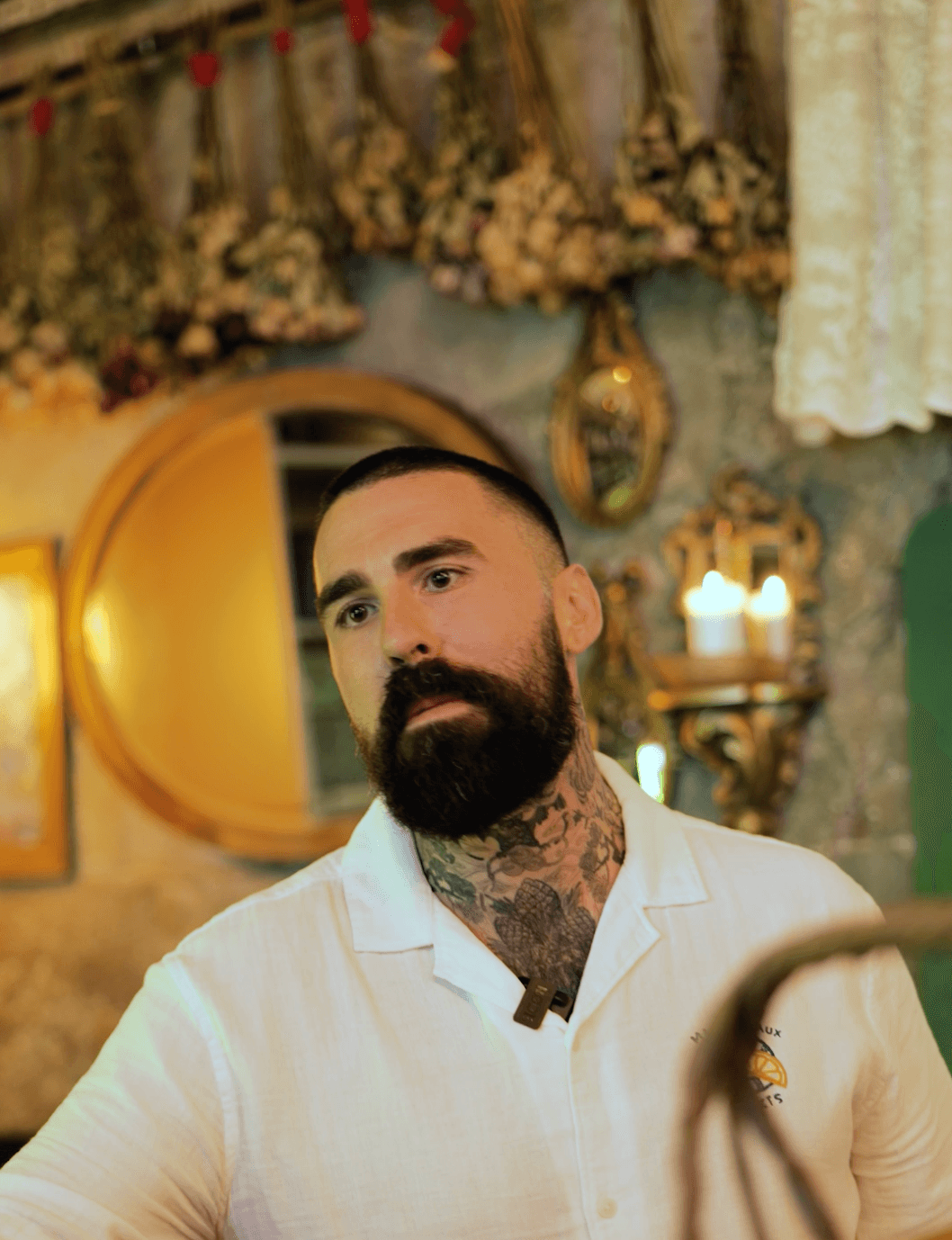 Portrait of a bearded man with neck tattoos wearing a cream-coloured chef's jacket in an ornate restaurant interior with decorative wall panels and hanging dried flowers.