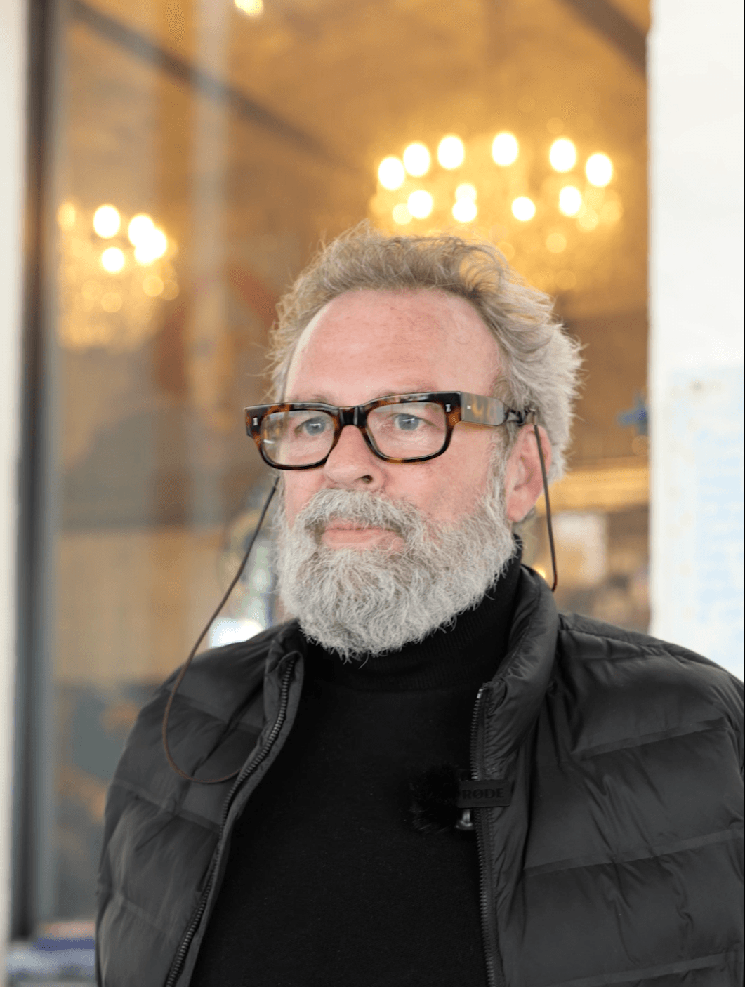 Professional portrait of an older man with grey hair and beard wearing glasses and a black puffer jacket, photographed against a blurred background with warm lighting.