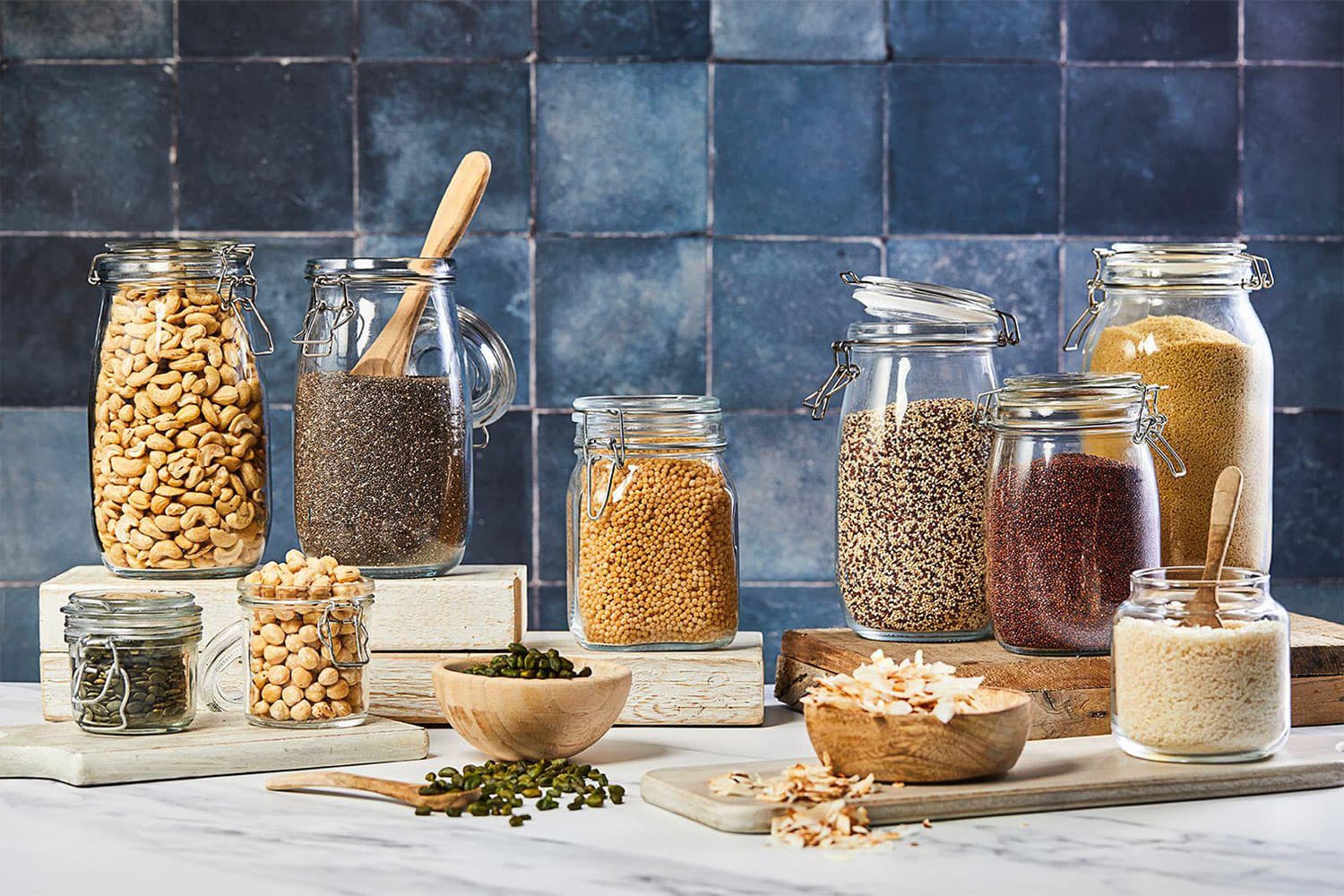 Collection of glass storage jars with airtight lids containing various dried ingredients including cashews, chia seeds, lentils, quinoa, and rice, arranged on wooden blocks against a blue tiled background with fresh herbs and wooden bowls.