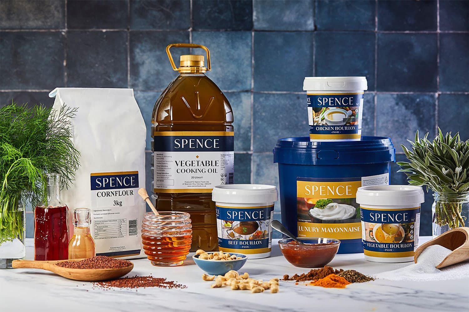 A styled product photography arrangement featuring Spence brand cooking ingredients and condiments including vegetable oil, cornflour, bouillons, mayonnaise, and spices, displayed with fresh herbs, grains, and cooking utensils against a blue tiled background.