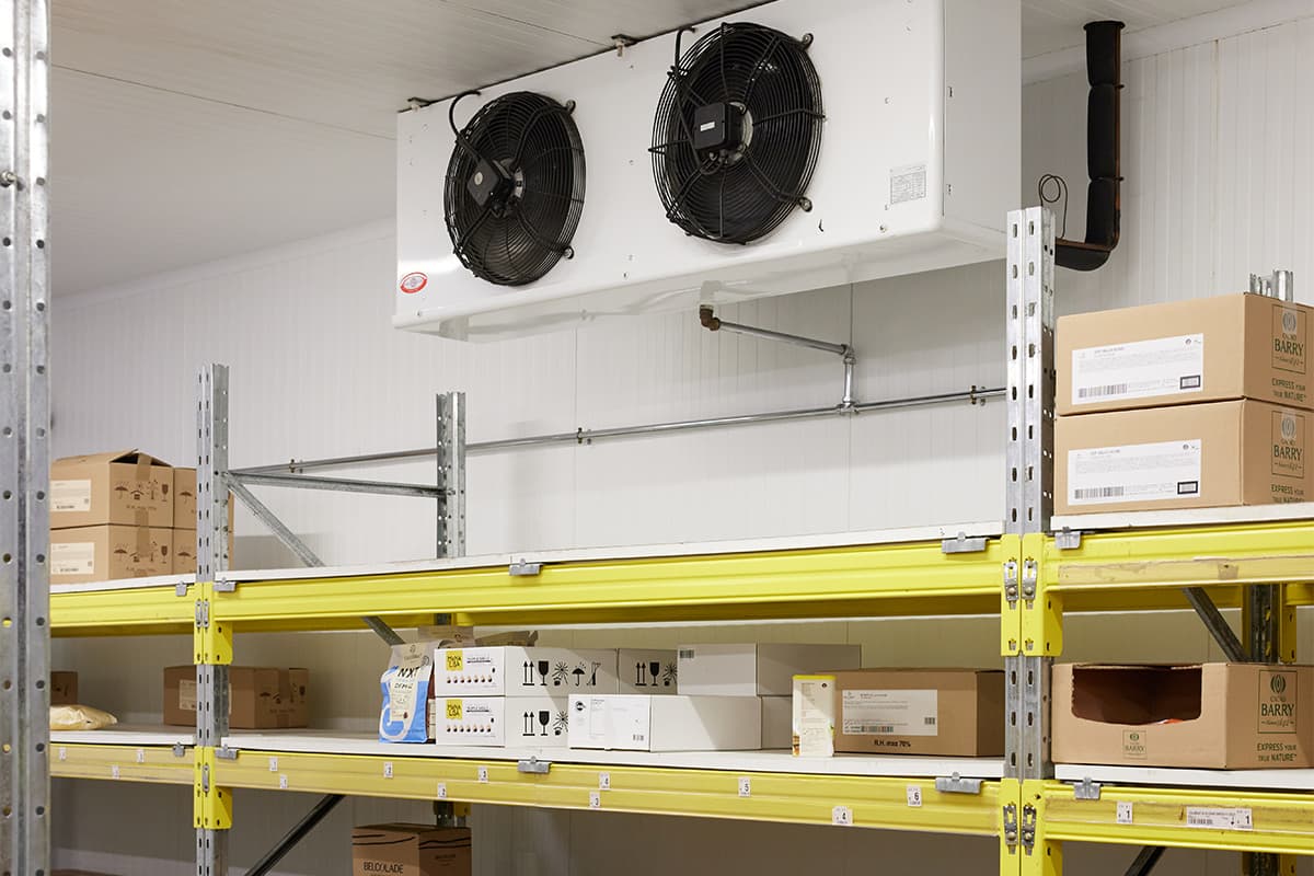 Interior view of a temperature-controlled warehouse storage room featuring industrial cooling fans mounted on the ceiling, metal shelving units with yellow beams, and cardboard boxes stored on multiple shelf levels.