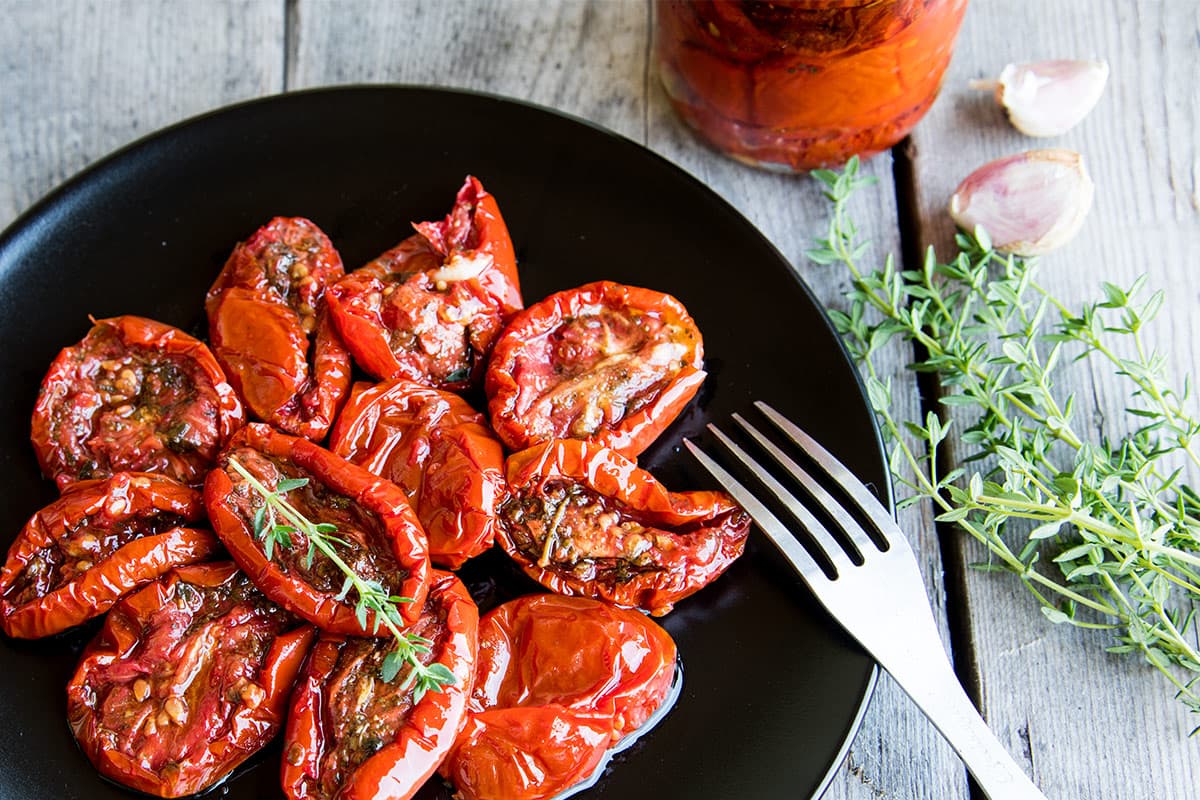 A black plate filled with roasted red peppers garnished with fresh rosemary, accompanied by a fork, garlic cloves, fresh herbs, and a glass of tomato sauce on a wooden surface.