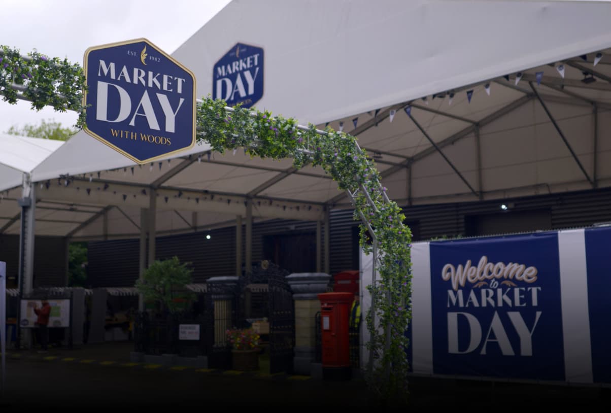 Exterior view of a market event venue with 'Market Day With Woods' branding, featuring covered stall areas, green ivy decoration, and a welcome banner.