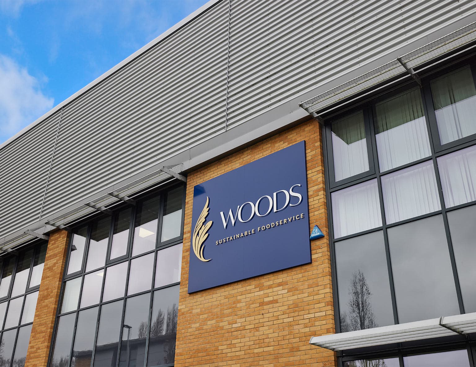 Exterior view of a modern commercial building with a blue signage panel displaying the Woods logo and 'Sustainable Foodservice' text mounted on a brick facade.