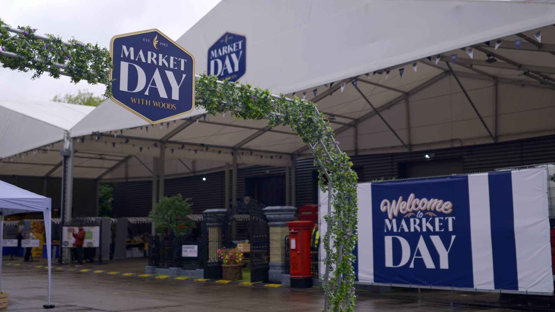 Covered market entrance decorated with 'Market Day With Woods' branding, featuring welcome banner, ivy garland, and event signage.