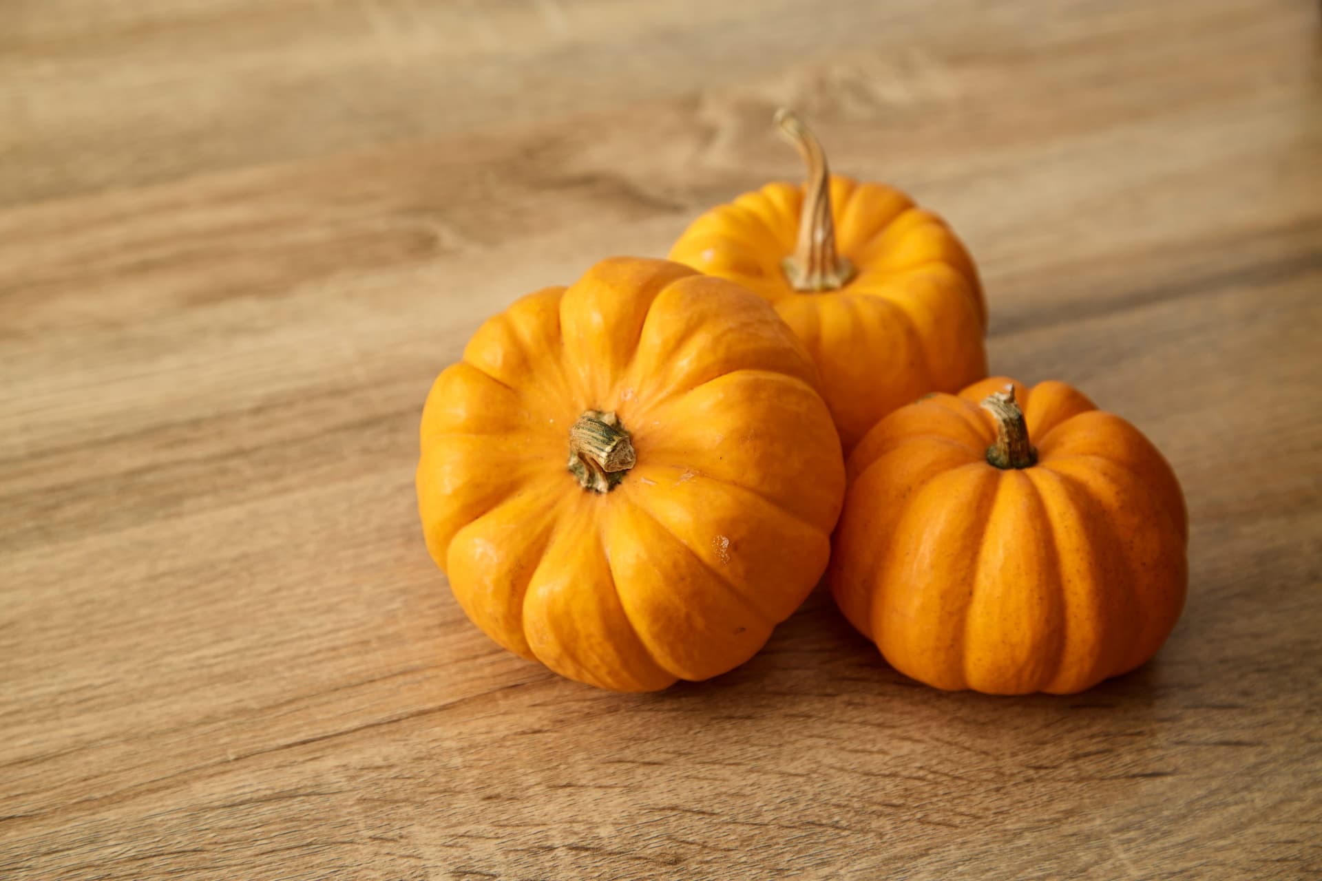 Three small orange pumpkins with stems arranged on a light wooden surface