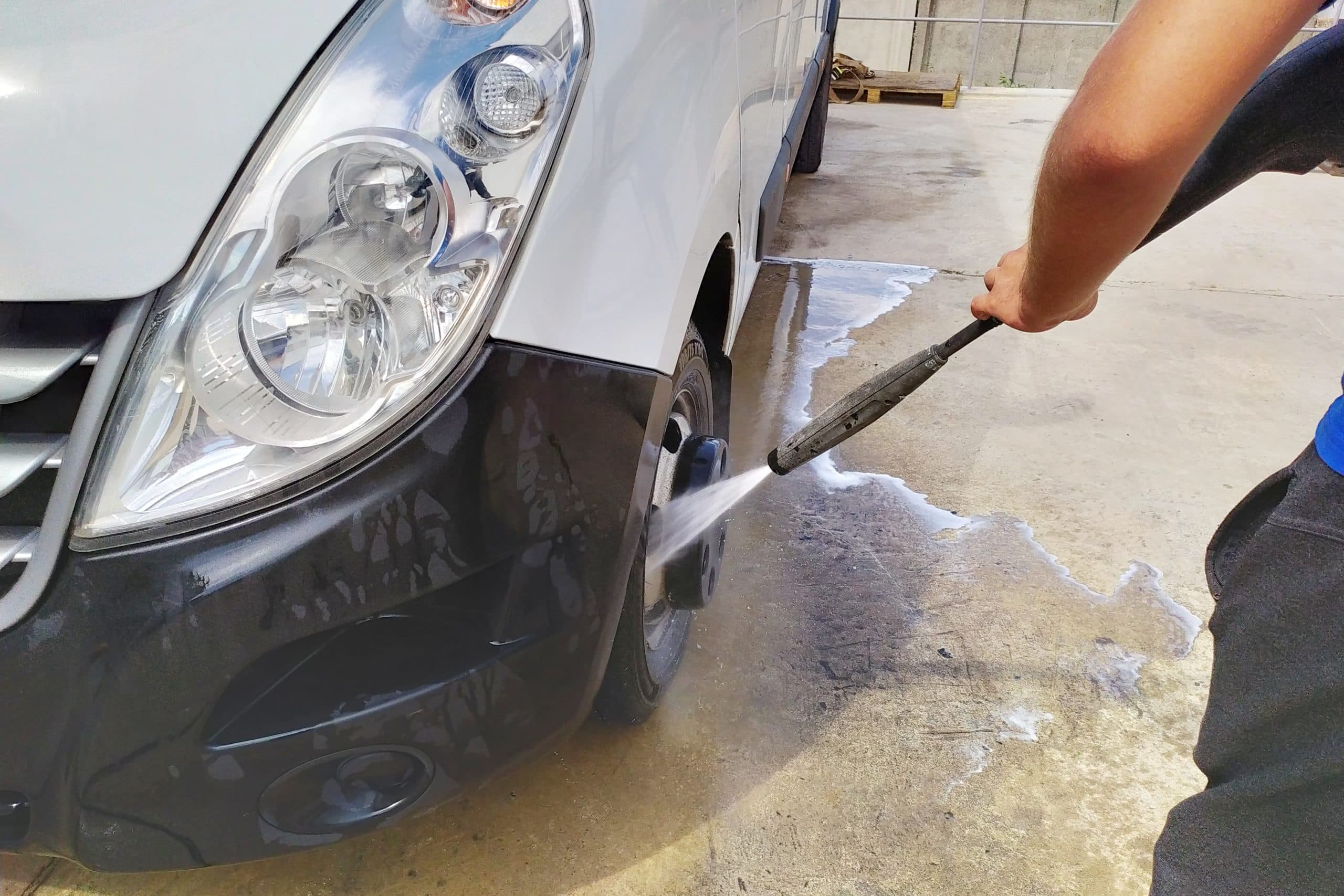A white car being cleaned with a pressure washer in a concrete warehouse or garage setting, with soapy water visible on the ground.