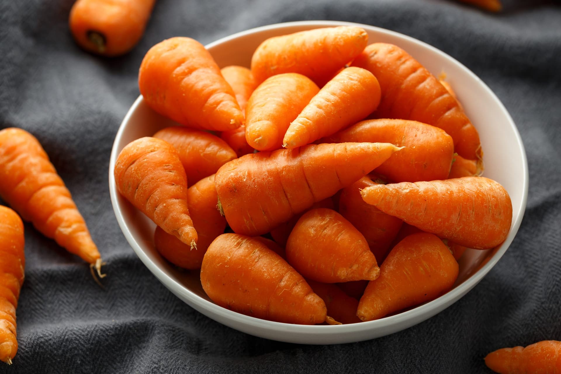 A white bowl filled with fresh orange carrots arranged on a dark grey fabric background, with additional whole carrots scattered around.