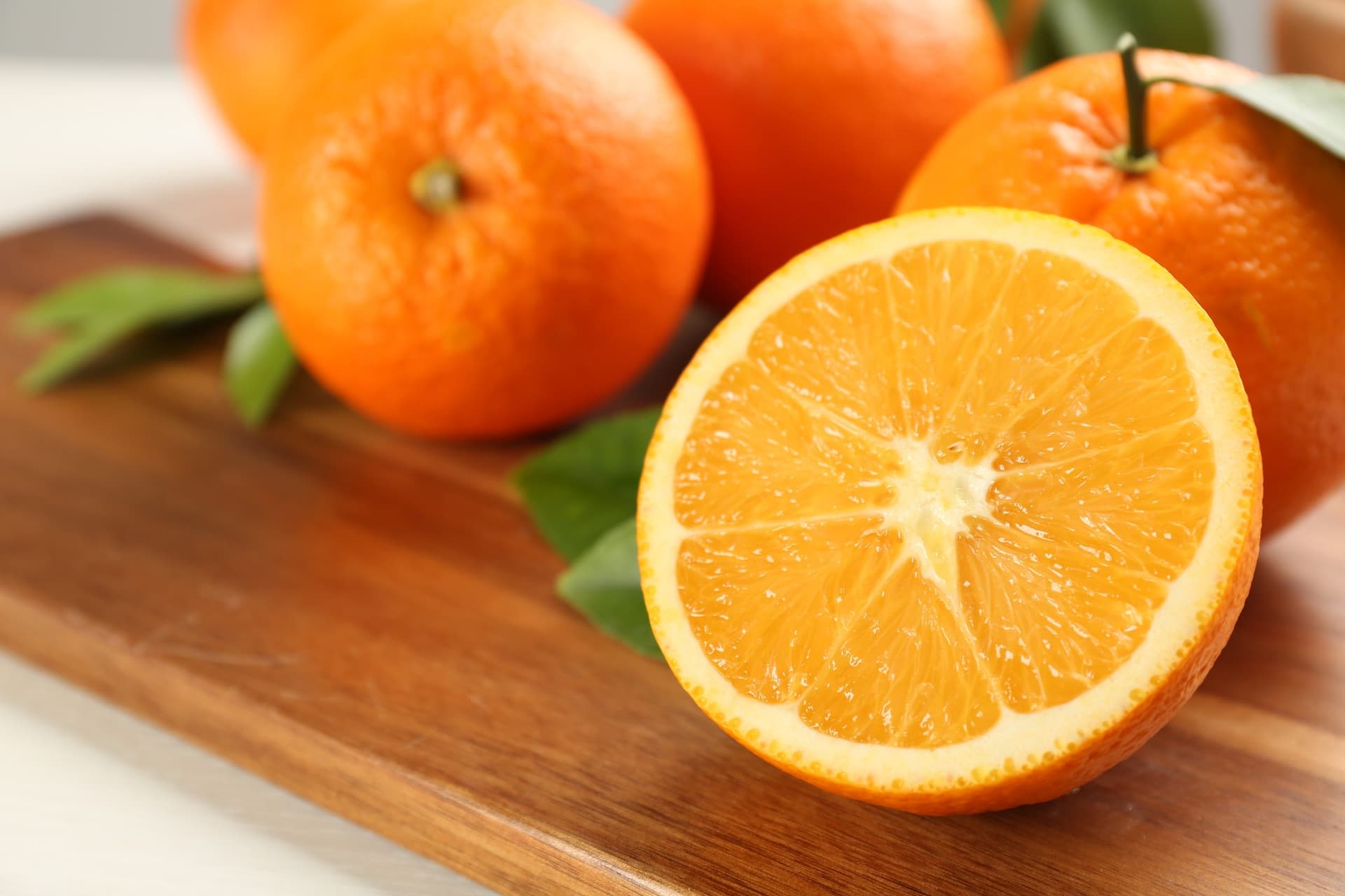 A halved orange displaying juicy segments on a wooden chopping board, surrounded by whole oranges and fresh green leaves.