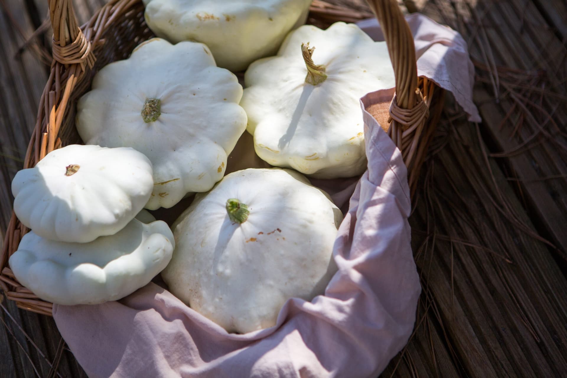 A wicker basket lined with purple cloth containing several freshly harvested white patty pan squash with green stems, photographed in natural sunlight.