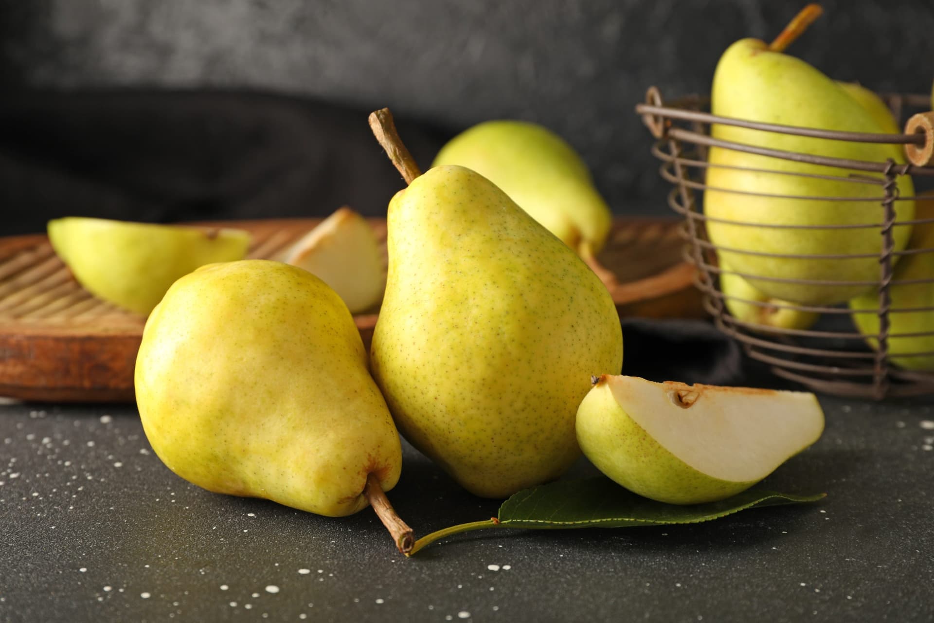 Three whole green pears and one sliced pear half displayed on a dark speckled stone surface, with a wire basket and wooden board in the background.