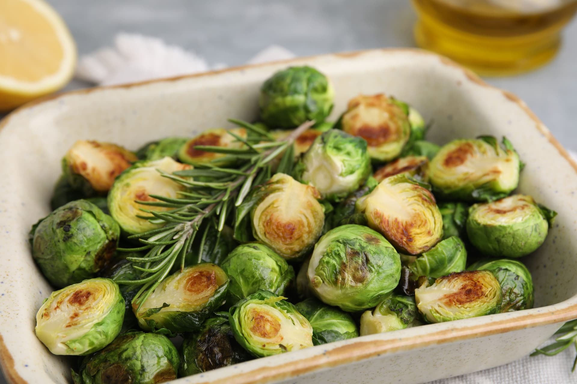A white serving dish filled with roasted Brussels sprouts halved and golden brown, garnished with fresh rosemary sprigs, with lemon and olive oil visible in the background.